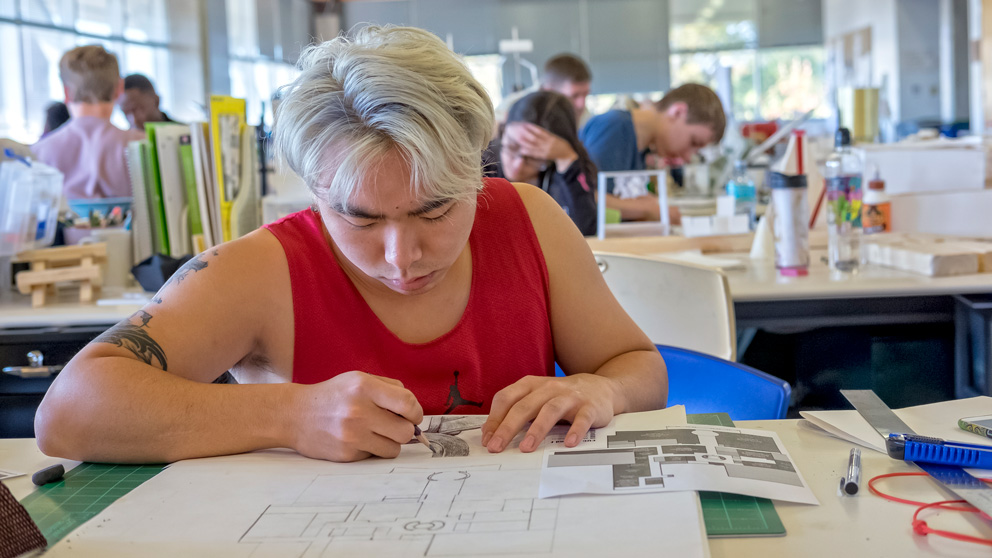 A student draws on paper at a workspace. Behind him, other students are working at desks.