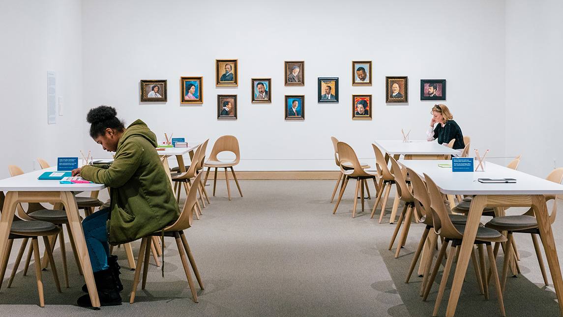 Two students study in a stark white gallery room in the Weatherspoon with 12 portraits hanging on the wall.