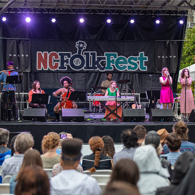 A band plays for an audience on a stage with a NC Folk Fest banner behind them.