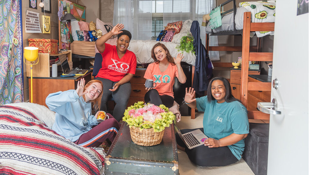 4 young women sit together in their college dorm room and wave to camera