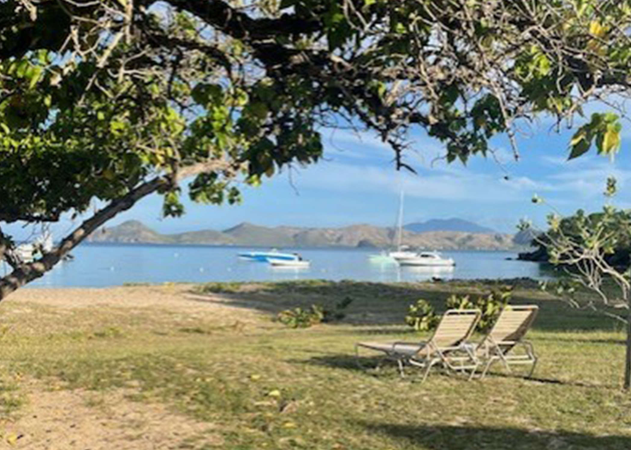 Carribean cove with sailboats in the distance and 2 chaise loungers in the foreground. 