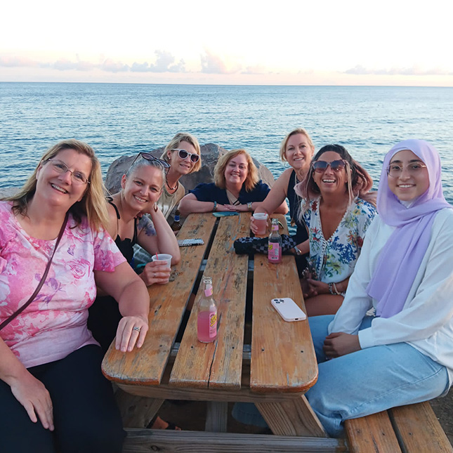 Seven women sitting at a picnic table with blue ocean behind them.