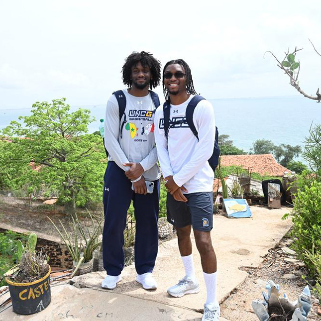 Two men wearing UNCG shirts and backpacks pose on a bluff overlooking the ocean with a thatched hut behind them.