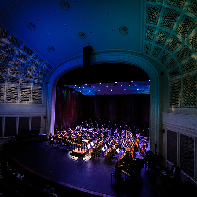 Wide shot of stage at UNCG Auditorium with a symphony and dark moody lighting.
