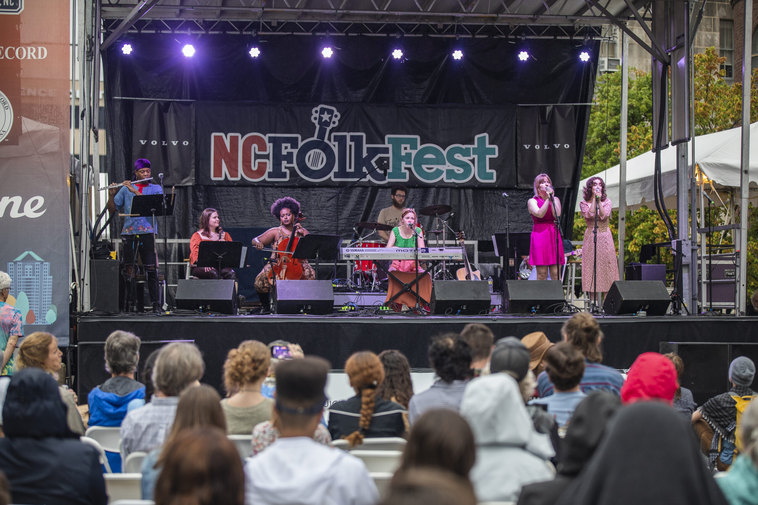 Band plays in front of a NC Folk Fest banner with filled audience seats in the foreground.