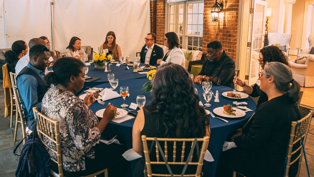 A large group of students and professors gather with UNCG Chancellor Gilliam at a dinner table.