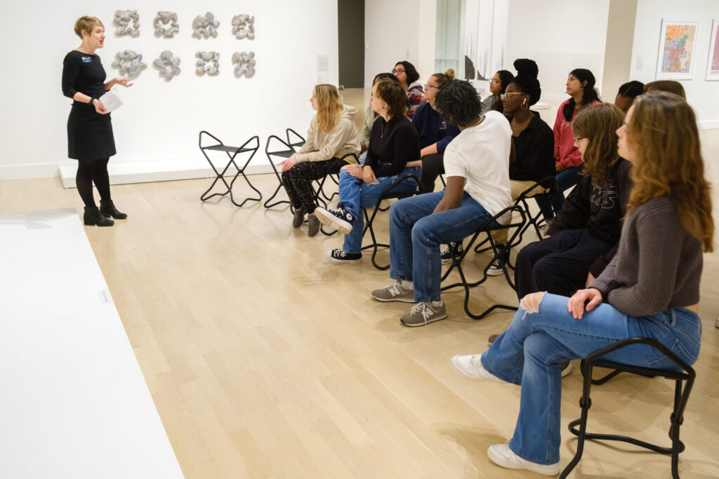 A woman leads a lecture in an art gallery as a group of seated students look on.