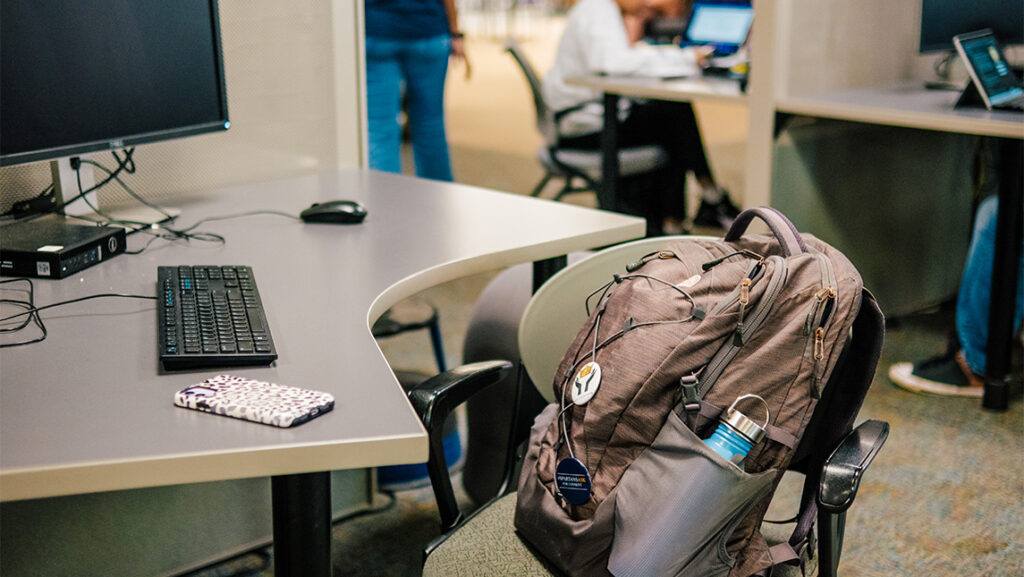 A backpack lies on a chair beside a computer.