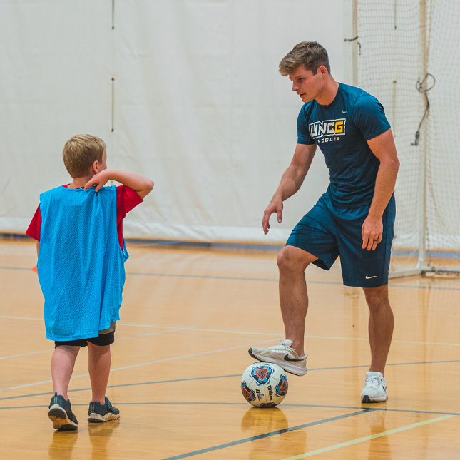 UNCG men's soccer player works with young boy in a gym.