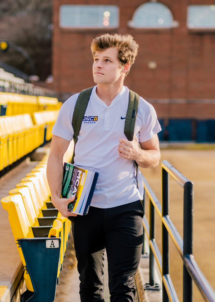 Man with UNCG soccer t-shirt walks in stands with a backpack and MCAT book.