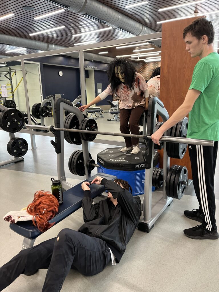 In a weight room, a student in a horror mask and make-up stands on a mat while another student lays on the floor underneath him to take a picture.