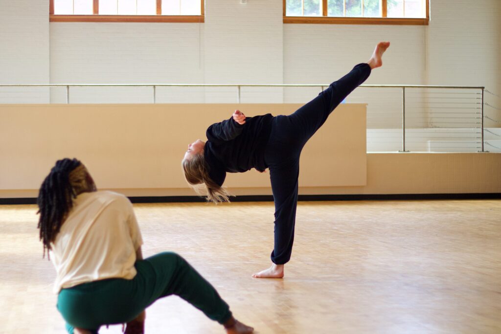 Two UNCG dance students practice a high kick move in a dance studio.