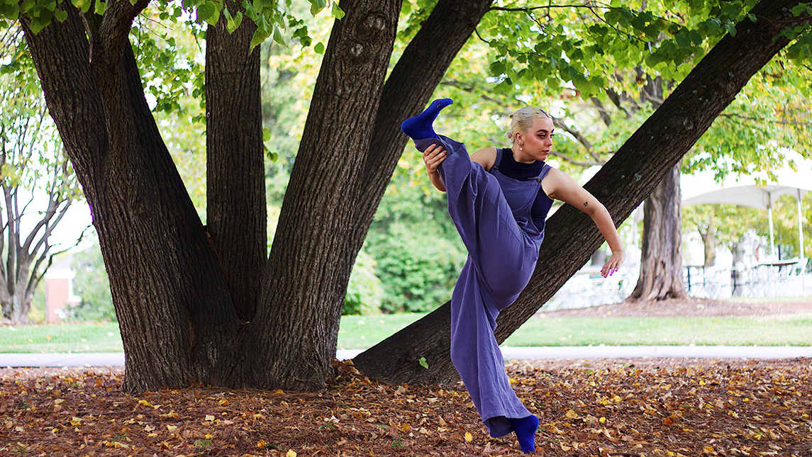 Dancer kicks her leg up as she practices outdoors in front of a big oak tree.