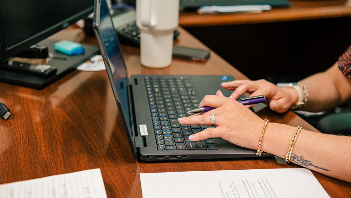 A close-up of a woman typing on a laptop at a UNCG office desk.
