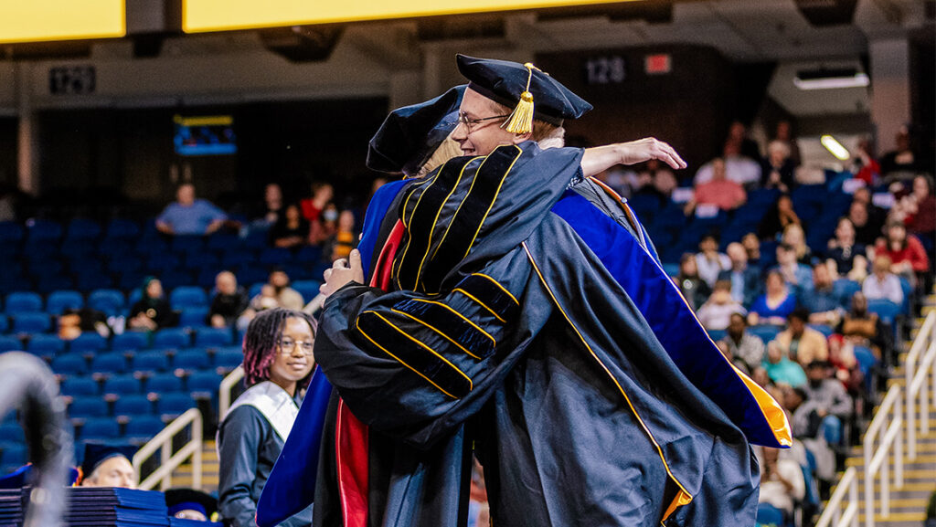 A graduating doctoral student hugs his mentor after being hooded at UNCG graduate ceremony.