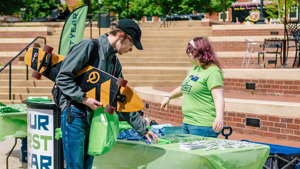 A UNCG student holding a skateboard talks to a Spartan SPEAR volunteer at a table outside.