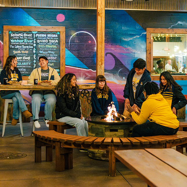 Students sit around a firepit in Southend Brewery. 2 more in the background sit at a bar eating apps.