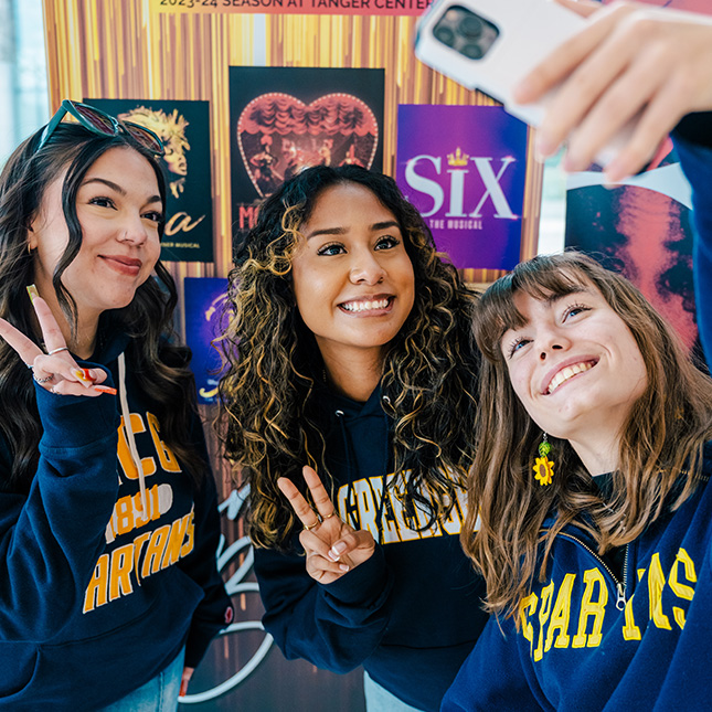 3 students in UNCG gear snap a selfie in the Tanger Center with posters of upcoming broadway shows in the background.