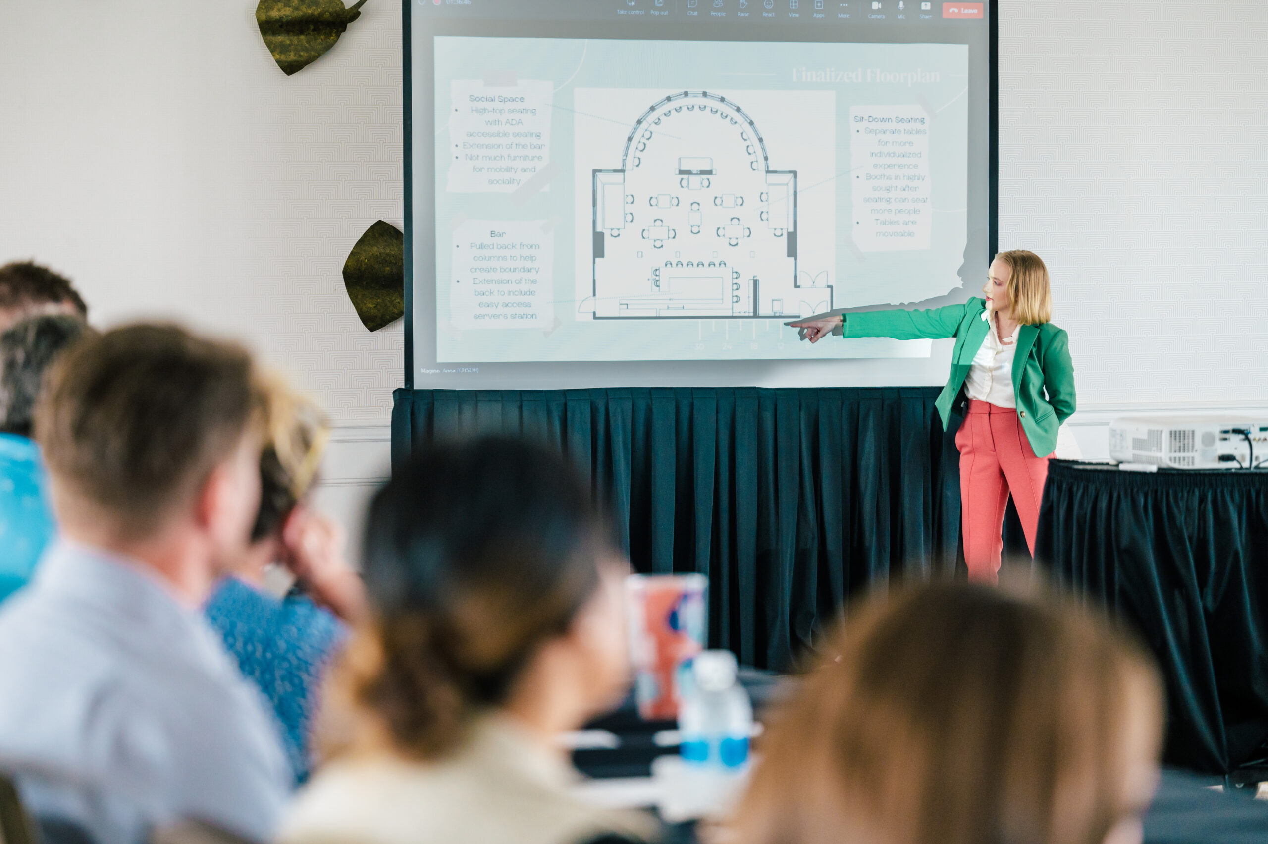 Student points to a floorplan on a AV screen in front of a group of professionals seated in a ballroom.