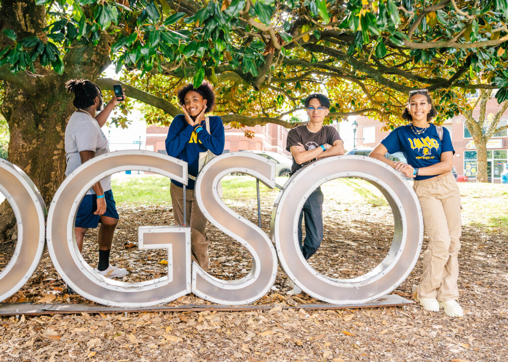 Students in UNCG gear pose with a GSO sign under a magnolia tree.