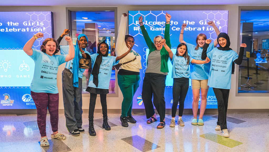 Six middle school girls in matching t-shirts jump and cheer with their two teachers in front of the "Girls Who Game Welcome" Monitor in the esports arena.