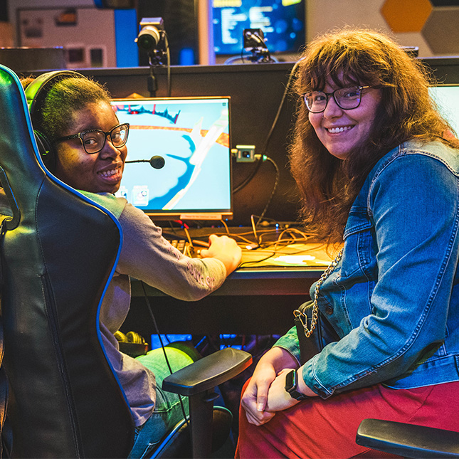 Two girls sit at the consoles in the esports arena and look over their shoulders to pose for the camera. 