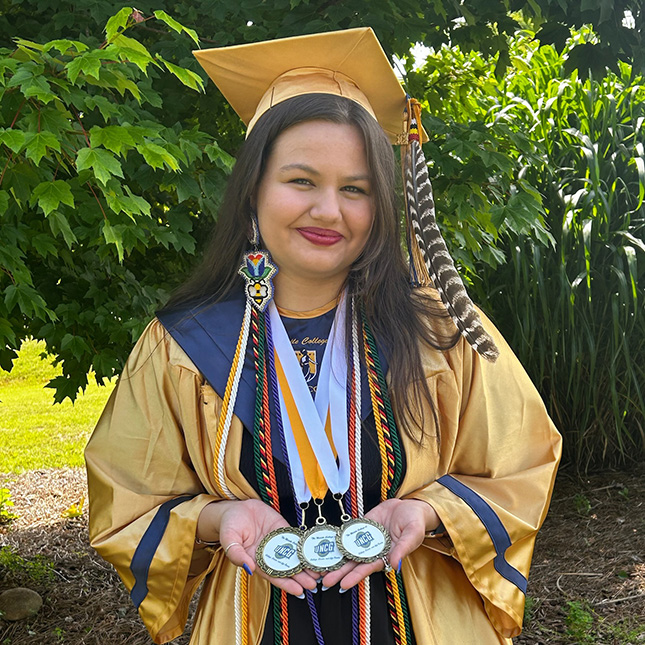Graduate poses in cap and gown with Native American accessories and many cords and medals to signify achievements.
