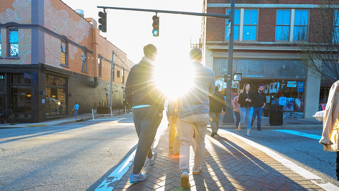 People are walking down the street in downtown Greensboro with the sun shining through.