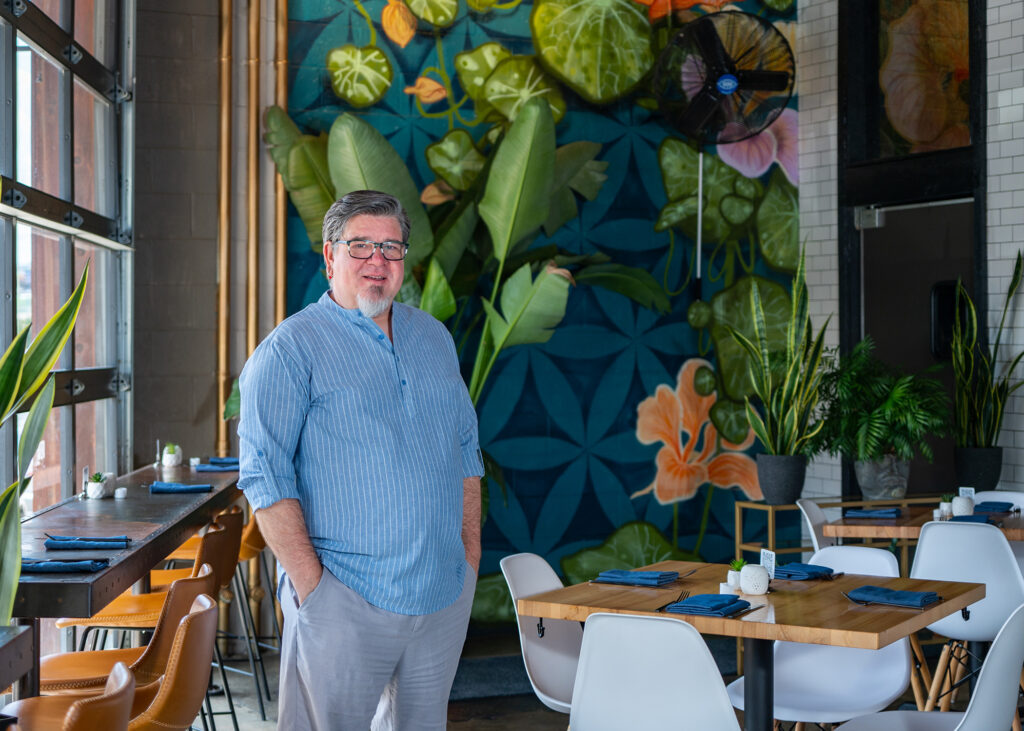 Man stands in a empty restaurant with a colorful mural of flora and fauna in the background and tables and chairs set for dinner.