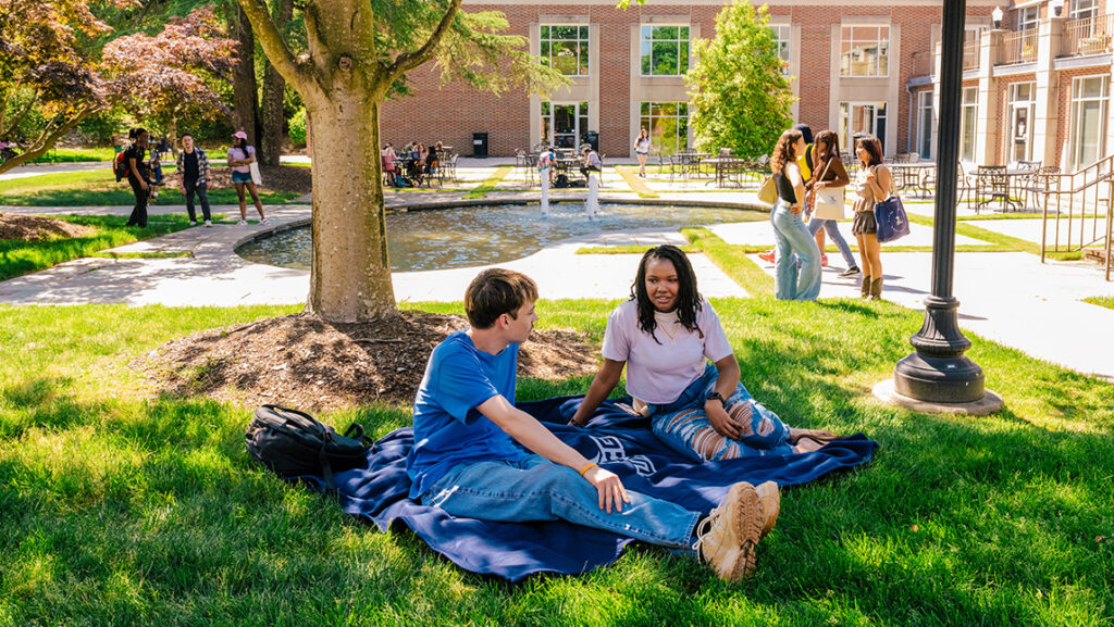 Two students sit on a blanket on campus with other groups talking behind them around the alumni house fountains.