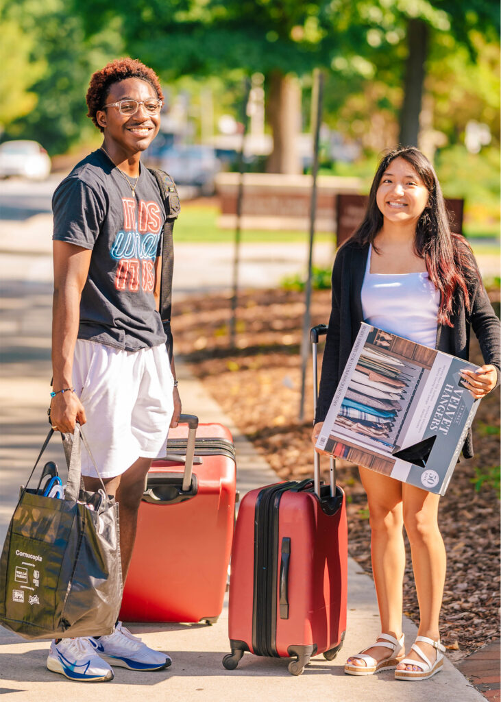 Two students smile as they roll suitcases and carry boxes down a campus sidewalk during move-in day.
