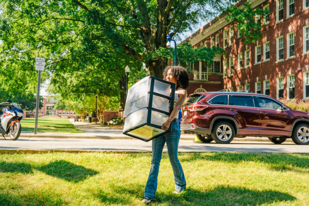 Student crosses lawn in front of residence hall carrying a plastic organizer.