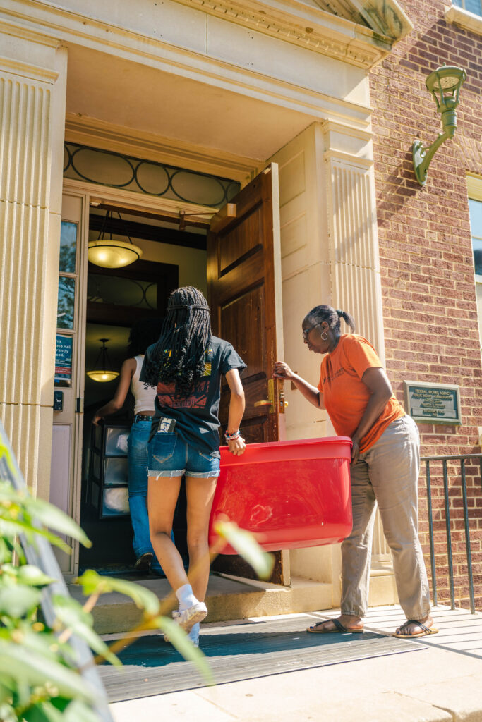 Mother opens the door to a residence hall as she helps her daughter carry a plastic bin inside during move-in.