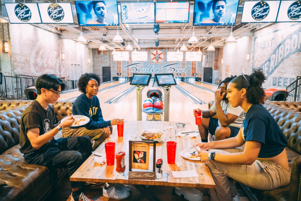 Four UNCG students eat at a table in Bourbon Bowl with the bowling lanes behind them.