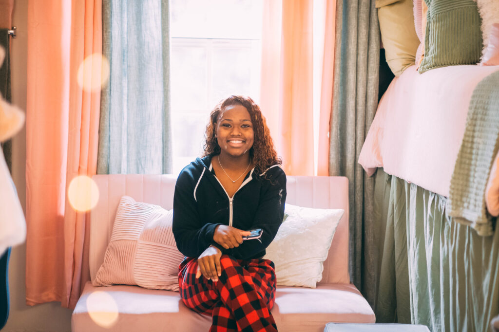 STudent sits on a love seat between two raised beds in her room with coordinated pink and green window treatments and bedding. 