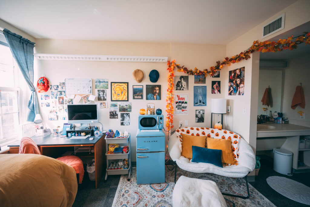 Residence hall room decorated with a blue retro mini fridge, a papasan chair, photos on the wall and a desk and bed with orange and blue decorations.
