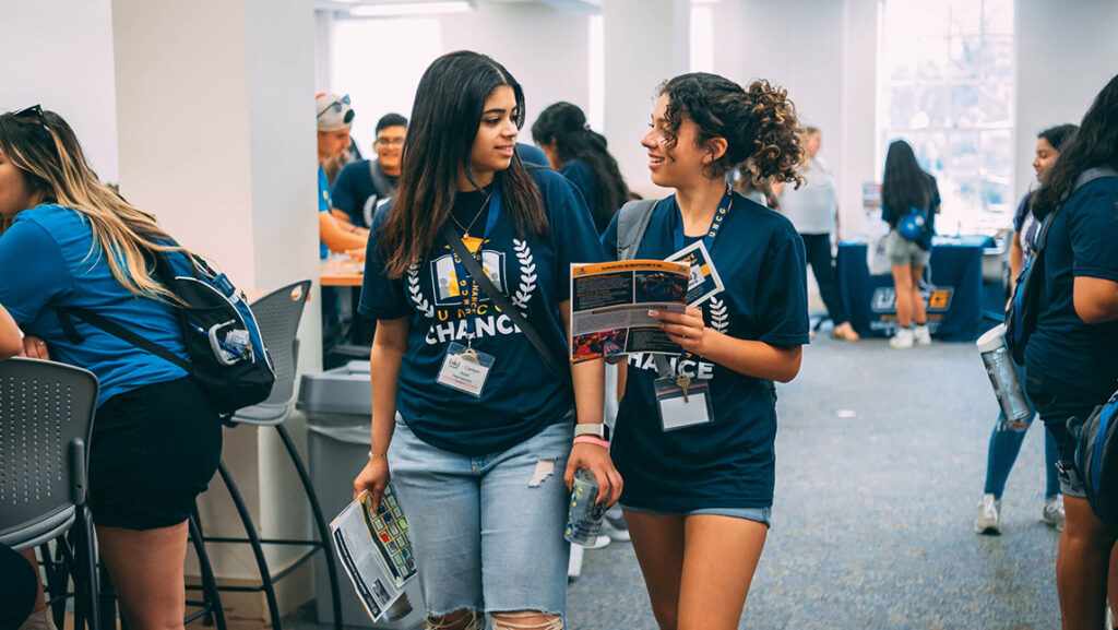 Two high school girls walk down a hall at UNCG while looking at a program.