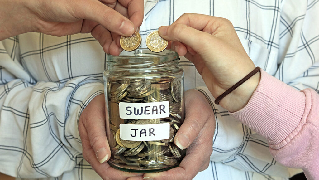 Close up on hands dropping coins into a jar with the label "Swear Jar."