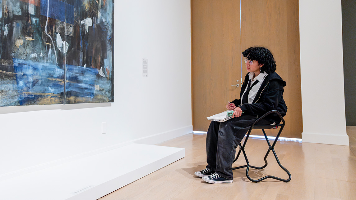 A UNCG student draws in a sketchbook while looking at a painting in the Weatherspoon Art Museum.