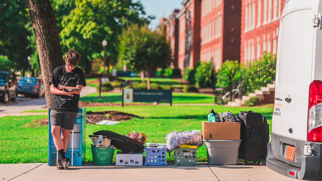 On a sunny day on the UNCG campus, a student leans against a box looking at a book with a line of boxes on a sidewalk and the back of a moving van on the right. Residence halls and green trees in the background.