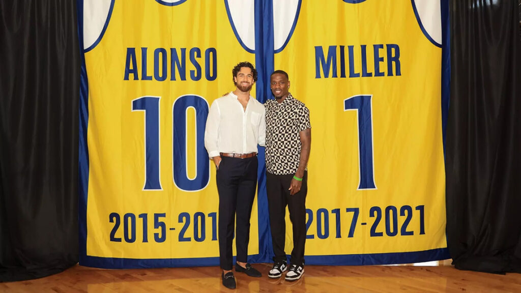 Basketball alumni Alonso and Miller in front of enlarged versions of their jerseys.