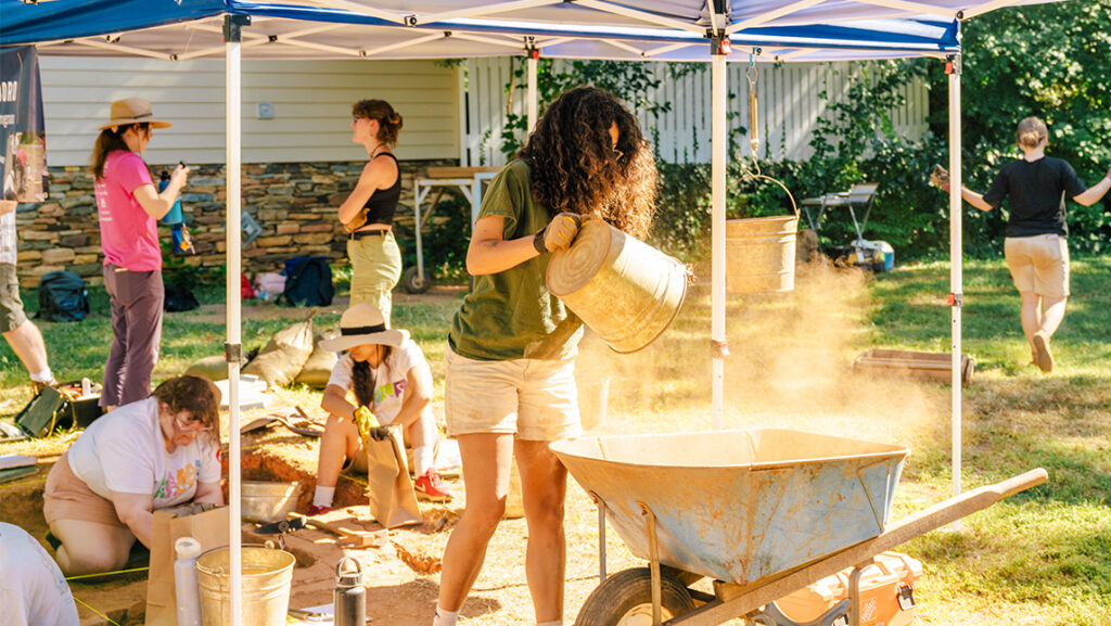 Young woman dumps sand from a bucket into a wheelbarrow as students behind her dig in a plot in the ground under a tent.