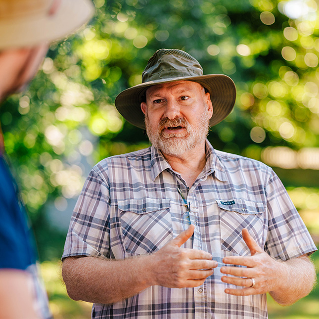 A man wearing a hat talks to another person using his hands outdoors.