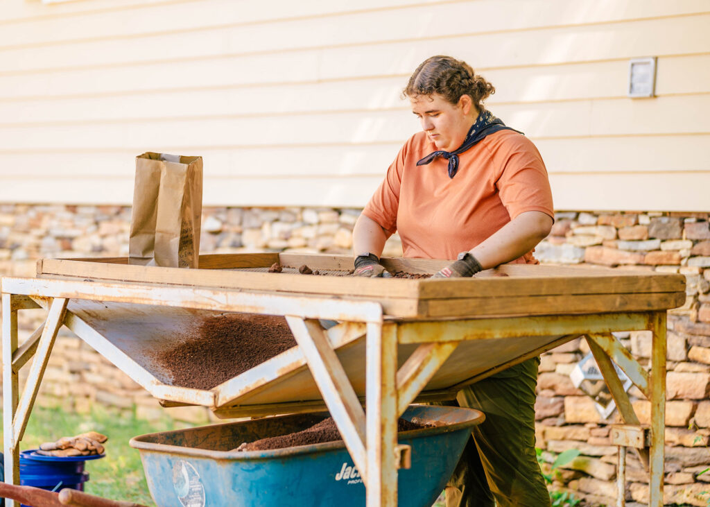 Woman stands at a table that sifts sand from heavier rock pieces. Sand falls into a wheelbarrow under the table.