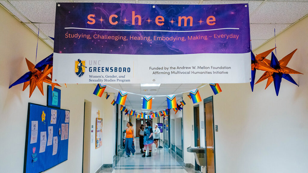 Banner hanging in a hallway with rainbow flags reads SCHEME. Down the hall we see students walking into a classroom.