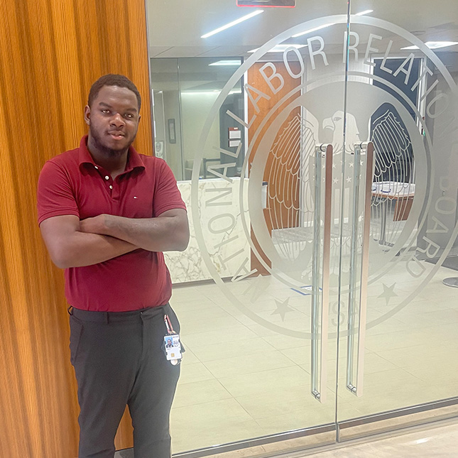 Young man stands with arms crossed in front of an office with glass doors etched with a seal for the National Labor Relations Board.
