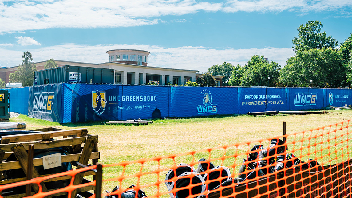 A construction fence with UNCG's EUC in the background.