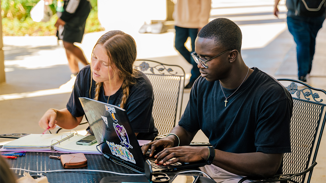 Two UNCG students use their laptop and notebooks at a outdoor table to study.