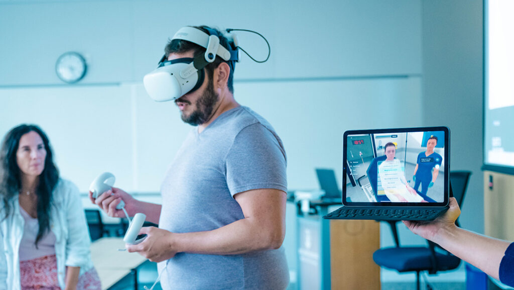 A UNCG student wears a virtual reality headset while someone holds up a laptop depicting a digital hospital room.