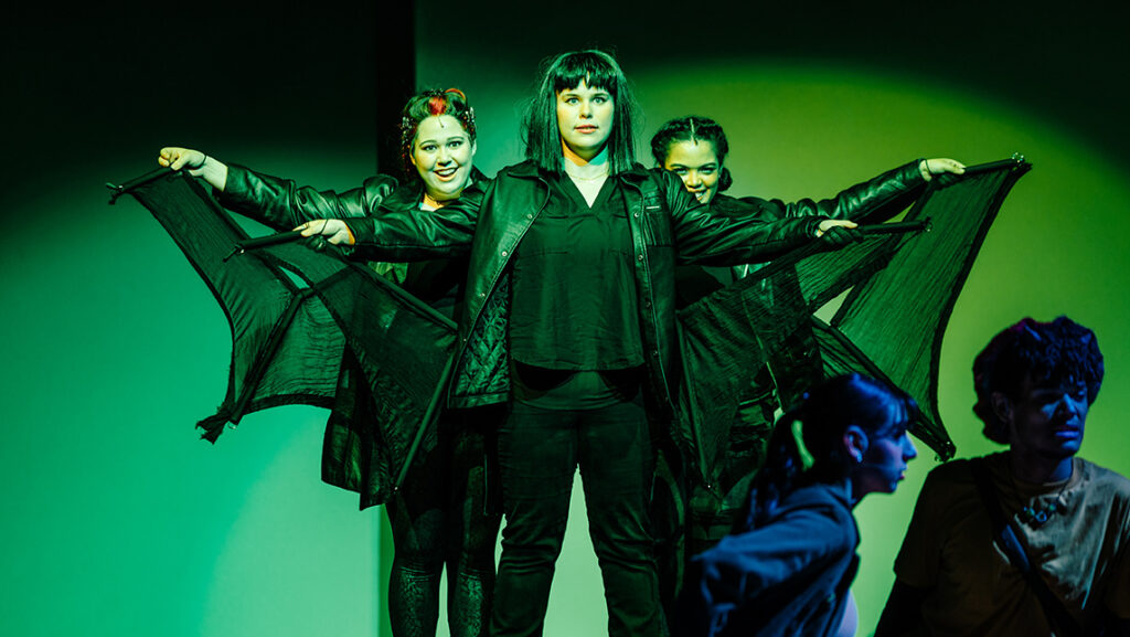 Three UNCG students stand onstage in black costumes with bat wings.